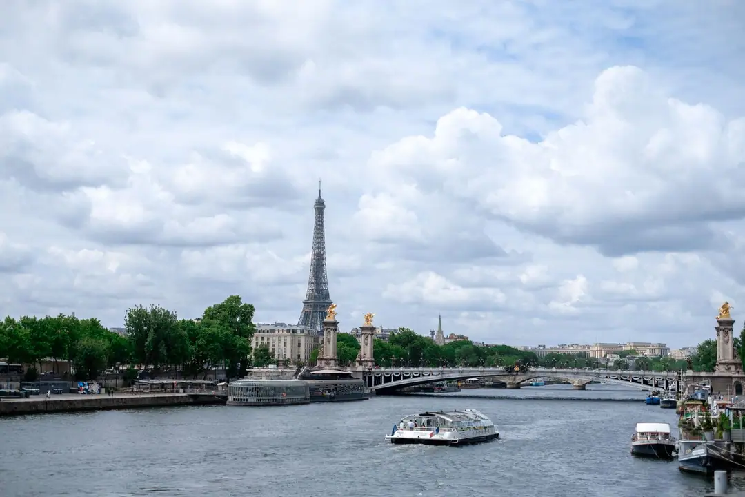 Scenic view of the Eiffel Tower and Seine River in Paris with a bridge and boats under a cloudy sky.